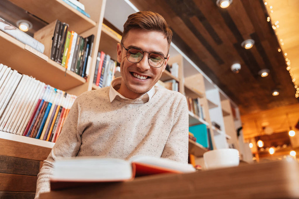 Young cheerful man sitting in cafe while reading book. - Photo, Image