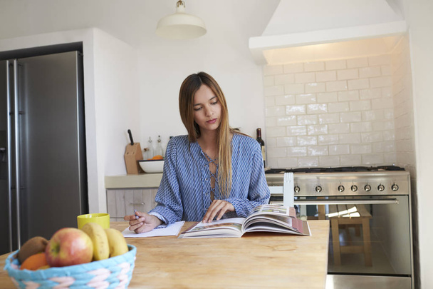 Woman sitting in kitchen and taking notes - Foto, Imagen