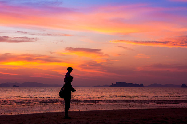Pai carregando seu filho no pescoço caminha na praia ao pôr do sol sobre o fundo do mar. estilo silhueta
 - Foto, Imagem