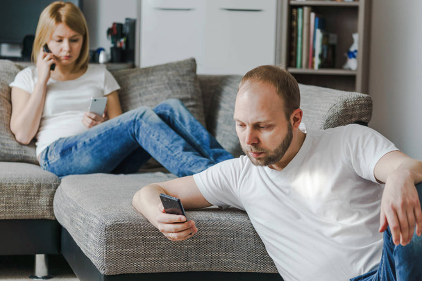 Νεαρή γυναίκα που κάθεται στο καναπέ μιλάμε στο τηλέφωνο, ενώ ο σύζυγός της χρησιμοποιώντας το κινητό του στο σαλόνι τους. - Φωτογραφία, εικόνα