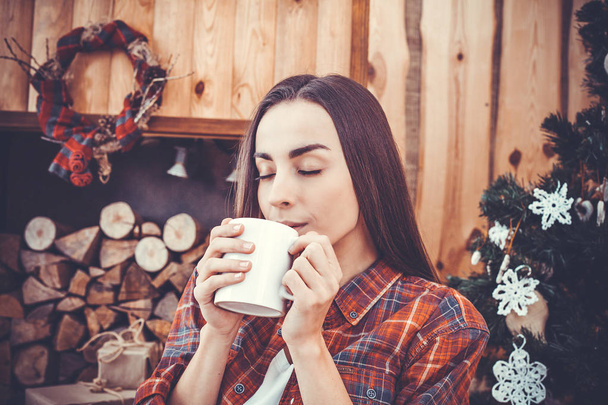 Portrait of a very beautiful and cheerful young girl the brunette on a background of the Christmas tree in a plaid shirt with red coffee mug in her hands. New Year concept. Girl smiling and resting. - Zdjęcie, obraz