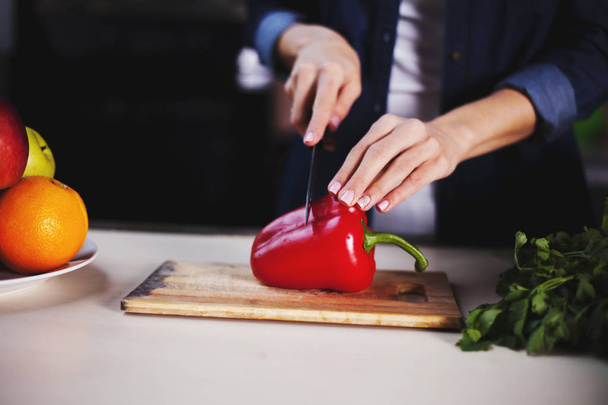 Young girl with a knife cuts red pepper at home in the kitchen on a wooden table. - Photo, image