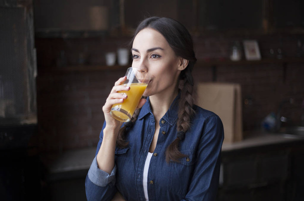 Portrait of very beautiful young brunette girl in jeans shirt on a kitchen at home. Girl laughing and holding orange juice in a transparent glass. - Foto, Imagem