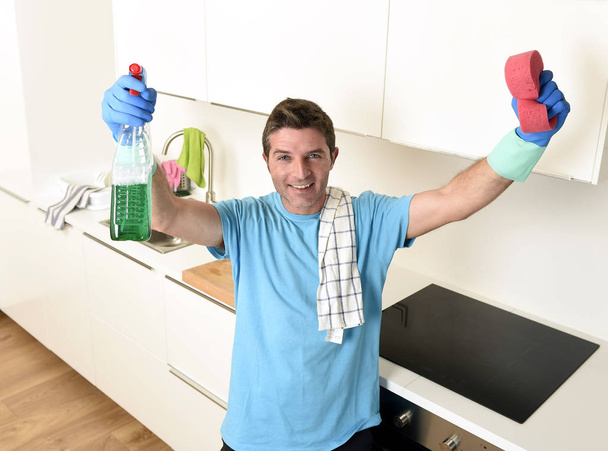młody człowiek szczęśliwy gospodarstwa prania detergentu sprayem i gąbka w rękawice uśmiechający się pewnie - Zdjęcie, obraz