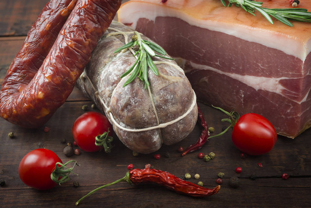 Italian salami, prosciutto ham and spicy sausage with cherry tomatoes and red pepper and spices close up on wooden background - Фото, изображение