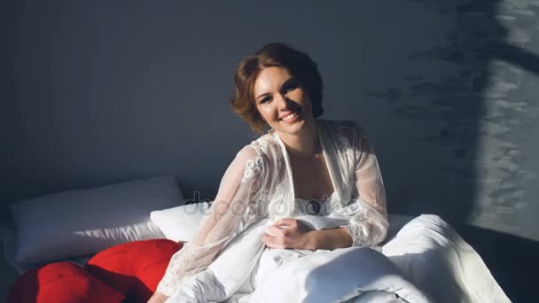 Portrait of beautiful young bride posing and smiling into camera Woman sits on the bed in bedroom - Filmati, video