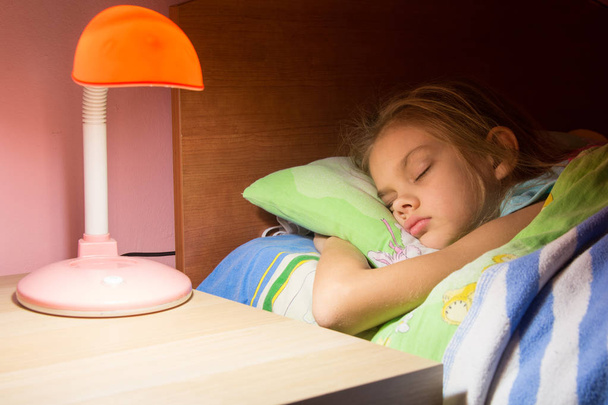 Семилетняя девочка спит в постели, чтение лампы включено на следующий стол
 - Фото, изображение