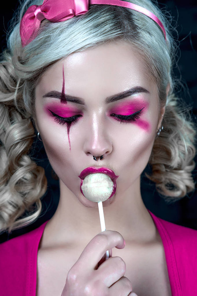 Mooie blonde meisje met twee pigtails, met creatieve pop make-up: roze glanzend lippen, het dragen van roze skelet jurk likken lollipop. voor de Halloween-party. Close-up, geretoucheerde foto. - Foto, afbeelding