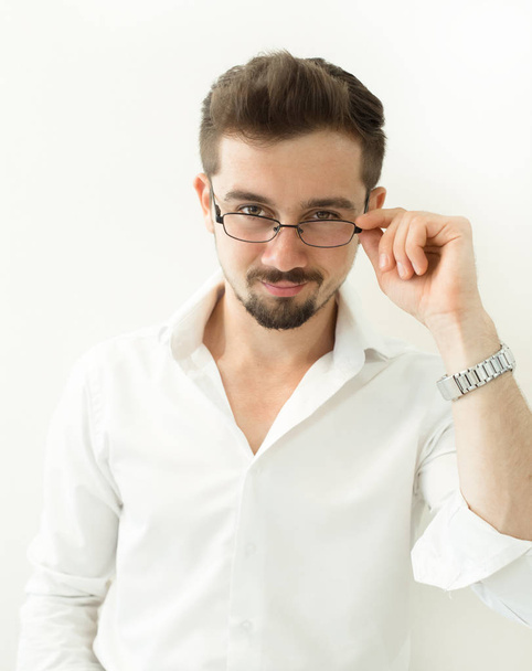 Όμορφος, έξυπνος άνθρωπος εκμετάλλευση γυαλιά σταθεί ενάντια σε λευκό φόντο. Γέρνει ο άνθρωπος σε γυαλιά και το άσπρο πουκάμισο, το πορτρέτο. Εσωτερικη χαμογελώντας νέος επιχειρηματίας φορώντας γυαλιά. - Φωτογραφία, εικόνα