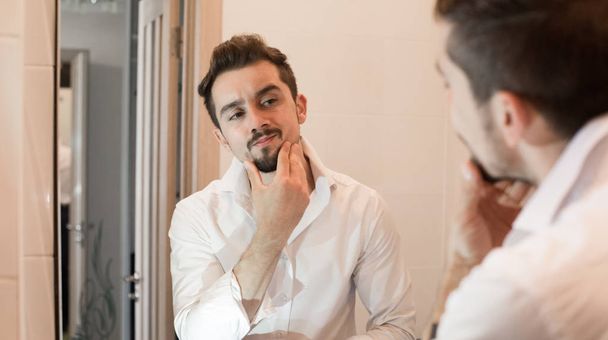 Man take care about his skin. Cheerful beaded guy caring of his skin. Handsome man look in mirror in bathroom in early morning - Photo, Image