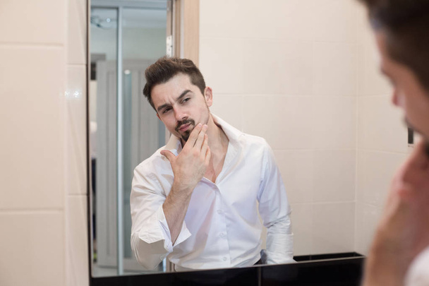 Man take care about his skin. Cheerful beaded guy caring of his skin. Handsome man look in mirror in bathroom in early morning - Foto, afbeelding