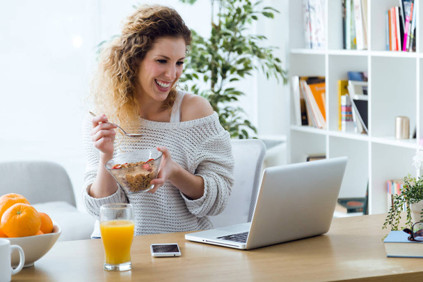 Beautiful young woman working with laptop while eating at home. - 写真・画像