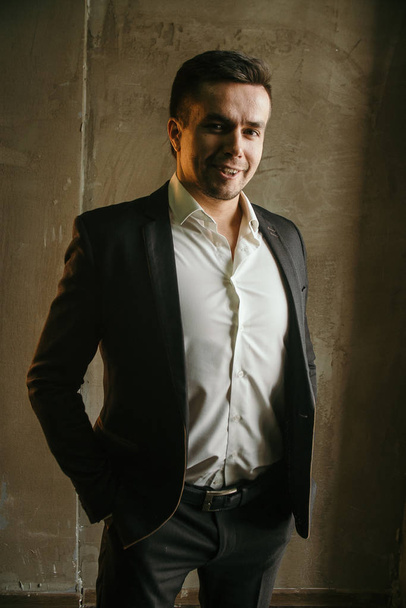 retrato de hombre de negocios guapo en traje sonriendo y posando con las manos en bolsillos en habitación oscura
 - Foto, imagen