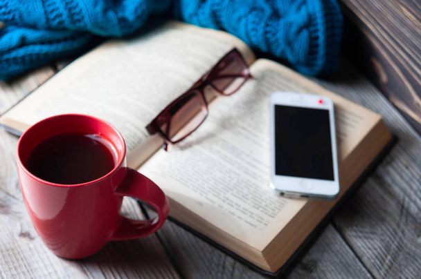 голубой уютный вязаный шарф с чашкой кофе или чая, телефоном, очками и открытой книгой на деревянном столе
. - Фото, изображение