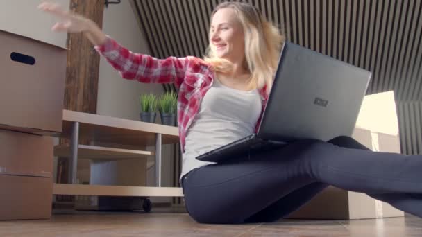 Beautiful woman is using a laptop excited smiling to find new stuff while sitting on the floor near the moving boxes - Video