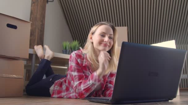 Beautiful woman is using a laptop excited smiling to find new stuff while sitting on the floor near the moving boxes - Filmagem, Vídeo