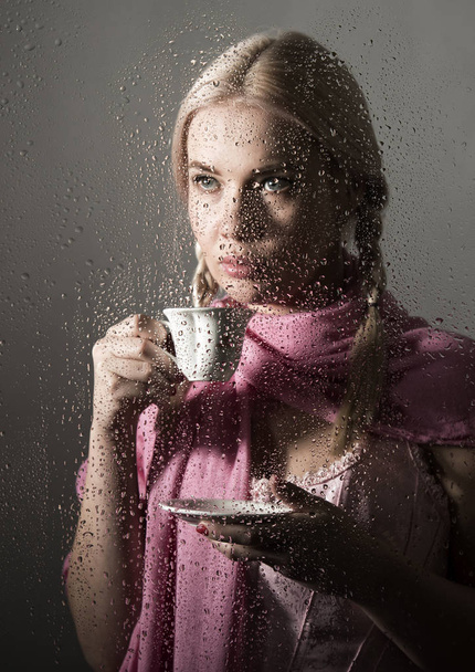 young woman dressed in sweater drinking coffee or tea, posing behind transparent glass covered by water drops - Foto, afbeelding