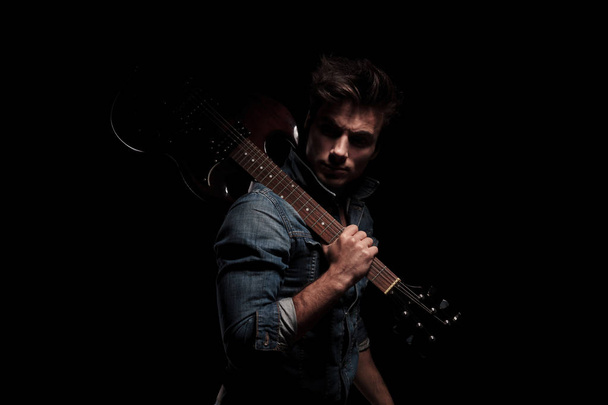 dramatické mladý kytarista, zatímco držení guitar sh při pohledu zpět - Fotografie, Obrázek