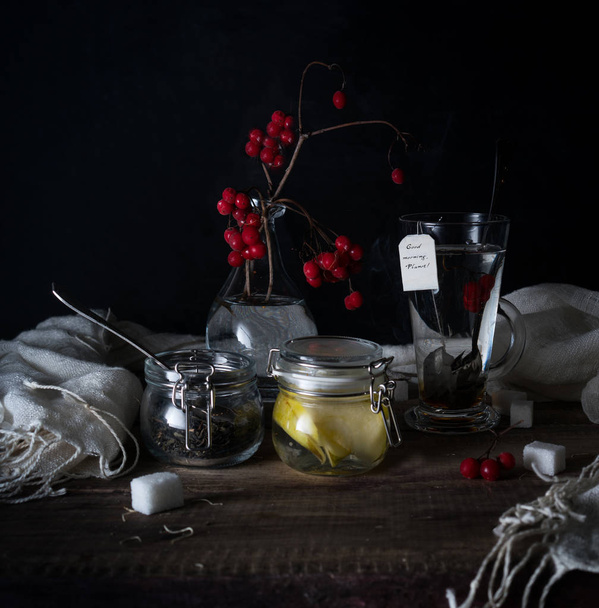 rustic still life. branch viburnum, tea in a big circle and jars on a wooden table. black background - Photo, Image