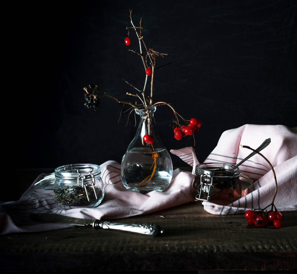 rustic still life. branch viburnum, tea in a big circle and jars on a wooden table. black background - Foto, afbeelding
