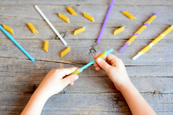 Pieni lapsi pitää olki ja kuivattu putki pastaa käsissään. Lapsi ripustaa pastaa pilliin. Yksinkertainen tehtävä hienomotoristen taitojen kehittämiseen lapsilla
 - Valokuva, kuva