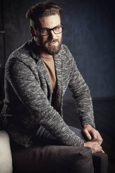 Portrait of handsome smiling trendy man wearing grey and glasses, with beard, sitting on dark furniture - Foto, imagen