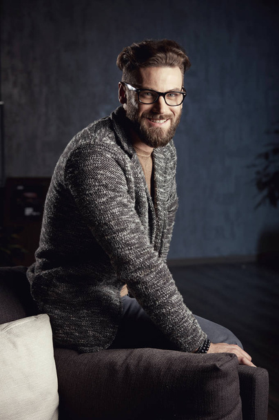Portrait of handsome serious elegant businessman wearing grey and glasses, with beard, sitting on luxury furniture in dark room - 写真・画像