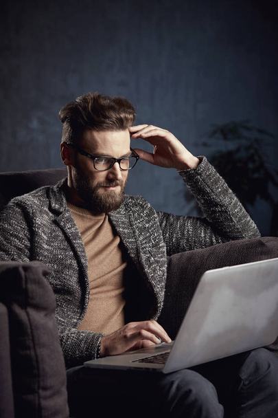 Attractive serious elegant modern businessman wearing grey and glasses, with beard, sitting on luxury furniture with laptop - Foto, Imagem