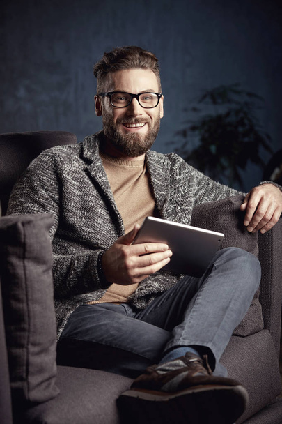 Attractive smiling elegant trendy man wearing grey and glasses, with beard, sitting on expensive sofa in fashionable dark interior  - Foto, afbeelding