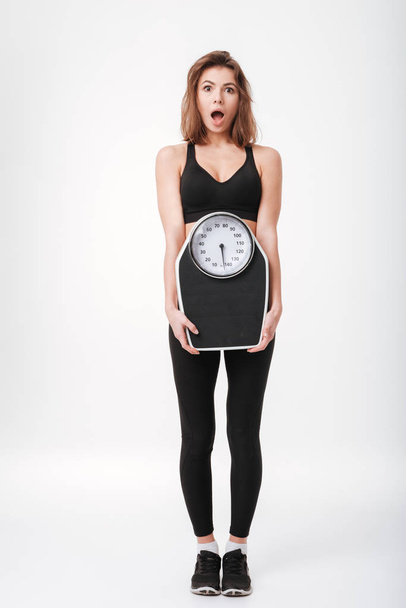 Shocked young fitness lady holding scales - Zdjęcie, obraz