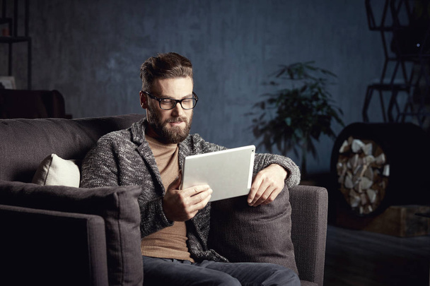 Handsome serious elegant classic man wearing trendy grey and glasses, with beard, sitting on stylish furniture in fashionable dark interior with laptop - Фото, изображение
