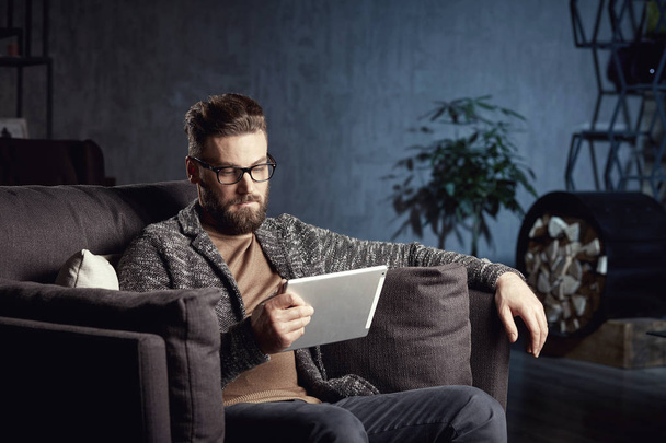 Handsome attractive elegant classic man wearing trendy grey and glasses, with beard, sitting on luxury furniture in fashionable dark interior with laptop - Foto, Imagen
