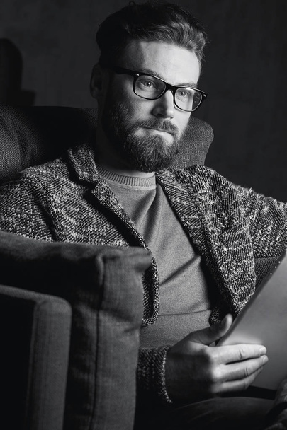 Portrait of handsome attractive elegant classic man wearing trendy grey and glasses, with beard, sitting on luxury furniture in fashionable dark interior with laptop in black and white - Foto, immagini