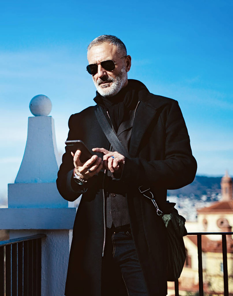 Pensive adult man wearing black aviator sunglasses and touching smartphone screen while spending time in city park.Vertical,blurred background. - Fotografie, Obrázek