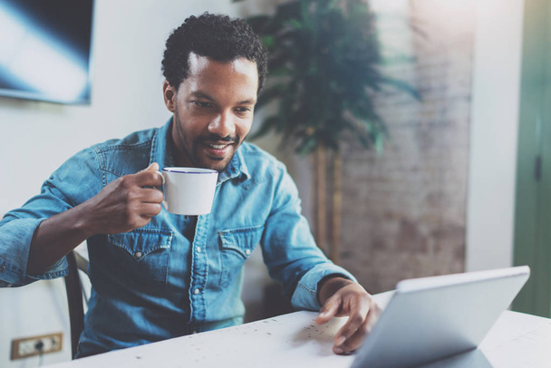 Smiling young African man making video conversation via digital tablet with friends while drinking black coffee in sunny office.Concept of happy business people.Blurred background, film effect. - Φωτογραφία, εικόνα