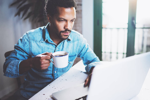Pensive bearded African man using laptop home while drinking cup black coffee at the wooden table.Concept of young people work mobile devices.Blurred window background. - Photo, image