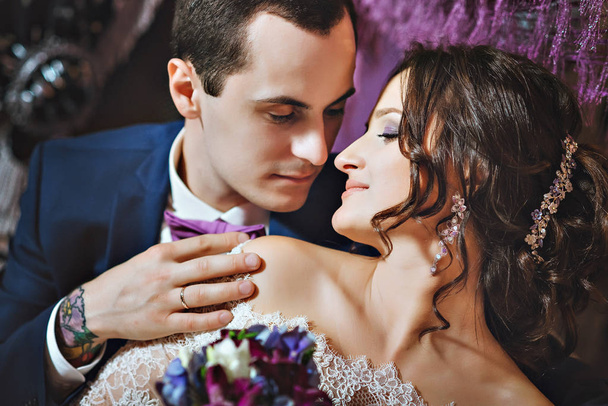 Την ημέρα του γάμου. Η νύφη και ο γαμπρός σε ένα πολυτελές εσωτερικό στο χρώμα της λεβάντας. Γάμος συναισθήματα. Όμορφη νύφη και κομψό γαμπρό στην τελετή. - Φωτογραφία, εικόνα
