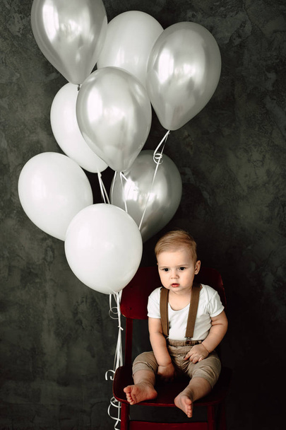 Портрет прекрасного маленького хлопчика щасливий усміхнений, що святкує 1 рік народження. Однорічний європейський маленький джентльмен сидить у кріслі і грає з повітряними кульками
 - Фото, зображення