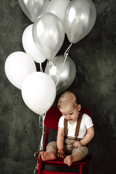 Retrato del adorable niño feliz sonriendo celebrando 1 año de cumpleaños. Un pequeño caballero europeo de un año sentado en una silla y jugando con globos
 - Foto, Imagen