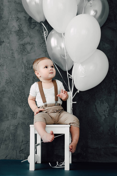 Porträt eines liebenswerten kleinen Jungen glücklich lächelnd, der 1 Jahr Geburtstag feiert. Ein Jahr alter europäischer kleiner Herr sitzt auf einem Stuhl und spielt mit Luftballons - Foto, Bild