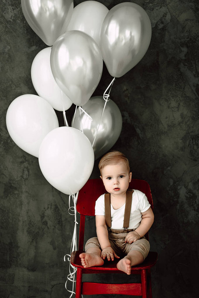 Muotokuva ihana pieni poika onnellinen hymyillen juhlii 1 vuosi syntymäpäivä. Vuoden vanha eurooppalainen pieni herrasmies istuu tuolissa ja leikkii balooneilla.
 - Valokuva, kuva