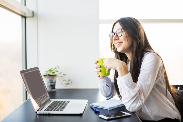 pracownik młoda kobieta siedzi w biurze podczas korzystania z laptopa na komputerze i pić kawy - Zdjęcie, obraz