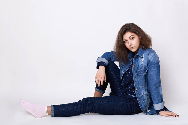 A cute girl wearing stylish jean clothes sitting on the floor indoors isolated over white background looking into camera with her charming dark eyes relaxing at home. Relaxed young woman in jeans - Fotó, kép