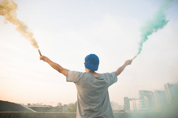 Male teenager holding colorful smoke sticks up in the air over urban city background - Photo, image