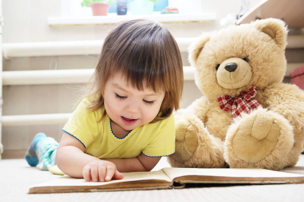 little girl reading book lying on stomach in her room on carpet with Teddy bear toy, smiling cute child, children education and development, happy childhood - Φωτογραφία, εικόνα