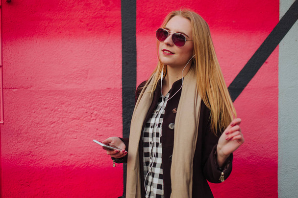 Nuori blondi tyttö aurinkolasit poseeraa lähellä värillinen seinä, kuulokkeet ja älykäs katsella
 - Valokuva, kuva
