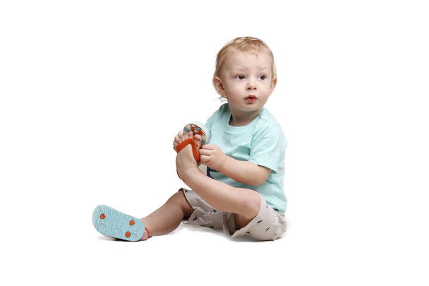 Маленький мальчик, сидящий на полу и держащий свою ногу изолированно над белым
 . - Фото, изображение