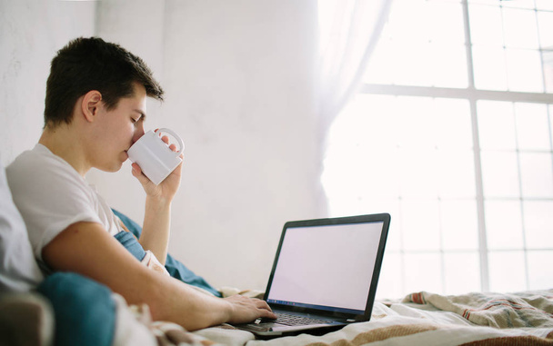 Rento nuori mies käyttää kannettavaa tietokonetta sängyssä kotona
 - Valokuva, kuva