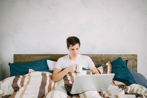 casual young man using laptop in bed at home and drinking coffee - Fotografie, Obrázek