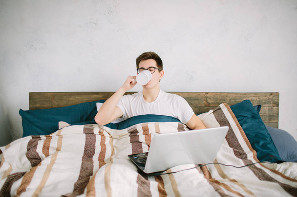 casual young man using laptop in bed at home and drinking coffee - 写真・画像